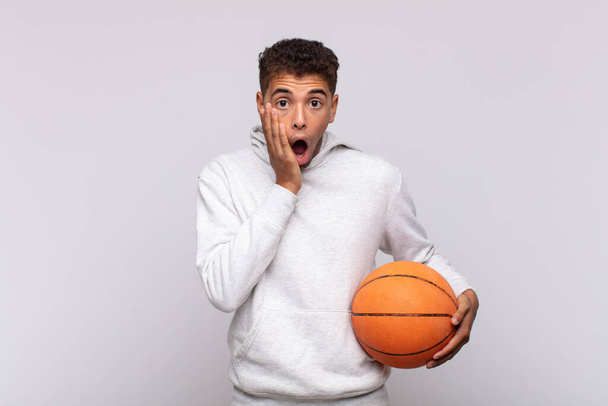 young man feeling shocked and scared, looking terrified with open mouth and hands on cheeks. basket concept - Foto, Imagem