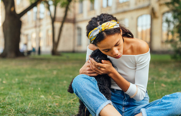 Bella donna che abbraccia un piccolo cane nero alleva un barboncino giocattolo seduto sull'erba nel parco. Tempo libero con un animale domestico all'aperto. - Foto, immagini