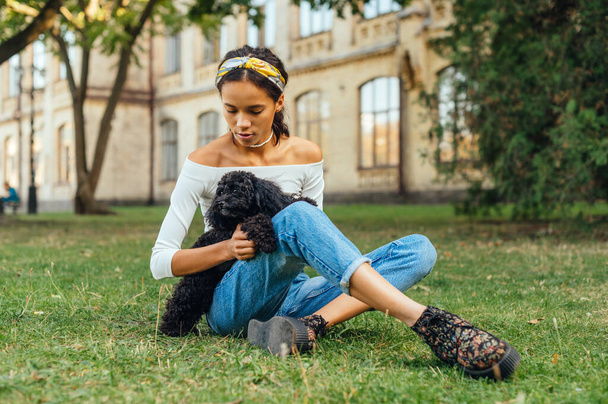 Porträt einer schönen hispanischen Frau, die auf dem Gras im Park vor einem architektonischen Hintergrund sitzt und mit einem kleinen schwarzen Hund auf dem Arm spielt. - Foto, Bild