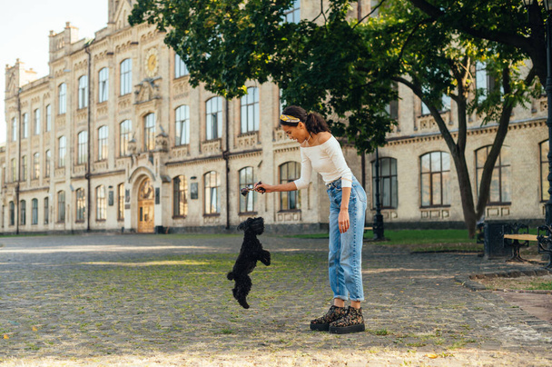 Femme mignonne en vêtements élégants jouant avec un chien dans le parc sur le fond de l'architecture ancienne, un petit chien noir saute dans les mains du propriétaire avec un jouet.. - Photo, image