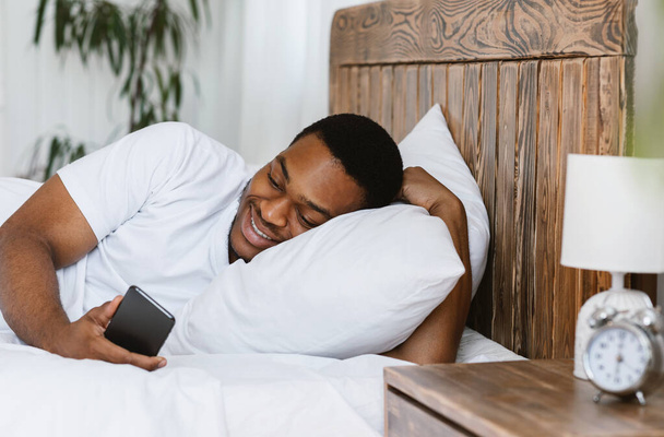 Счастливый африканский мужчина, пользующийся телефоном и просматривающий Интернет, лежа в кровати - Фото, изображение