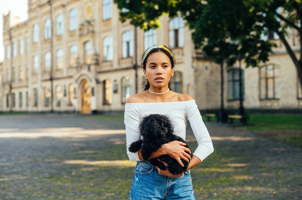Schöne Frau in stylischer Kleidung steht im Park und hält einen kleinen schwarzen Hund auf einem Hintergrund alter Architektur und schaut weg. Spaziergang mit einem kleinen schwarzen Hund. - Foto, Bild