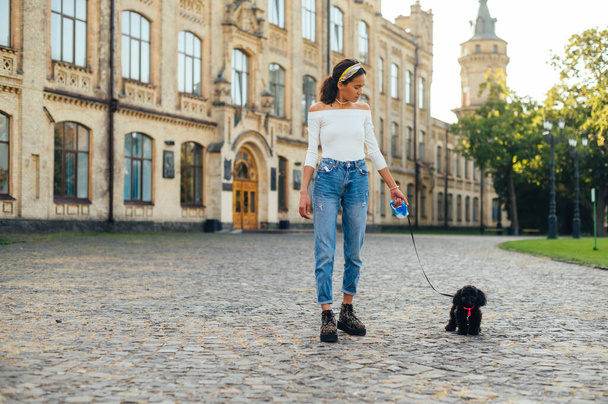 Stylish lady in casual clothes walks a small dog on a leash against the backdrop of a historic architect. Woman charred with a dog breed poodle toy. - Photo, Image