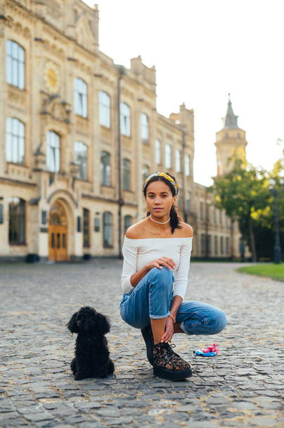 Attraktives hispanisches Mädchen, das mit einem Pudel aus der Hunderasse auf der Straße sitzt und für die Kamera posiert. Freizeit mit Hund - Foto, Bild