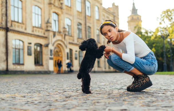 Stylish hispanic woman playing with a little black dog on the street with a serious face. Female owner on a walk with a dog on a background of historic architecture. - Photo, Image