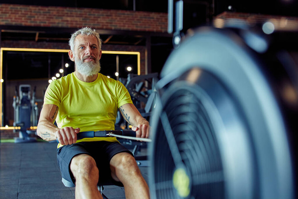 Entraînement cardio. Homme athlétique mature concentré fort en vêtements de sport faisant de l'exercice sur une machine à ramer au gymnase - Photo, image