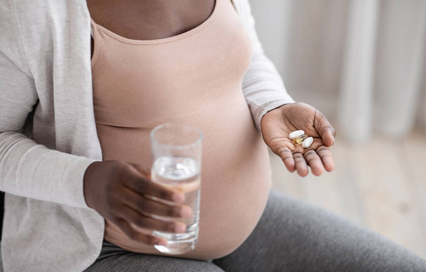 Vitaminas y suplementos prenatales. Mujer embarazada negra irreconocible que toma pastillas y agua - Foto, Imagen