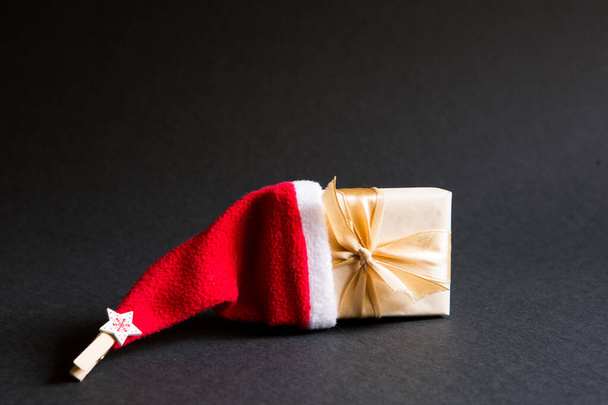A box with a Christmas gift in a package tied with a ribbon and with a Santa hat on a black background. Black Friday, buying gifts for the new year. Space for text - Photo, image