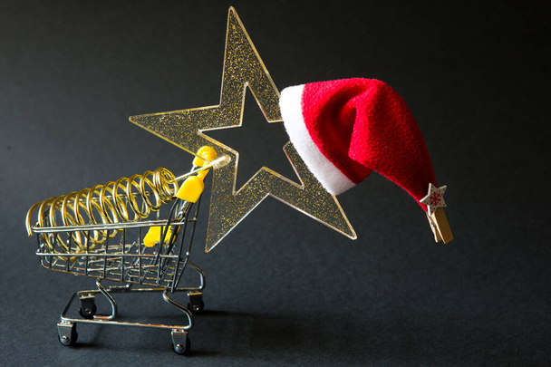 Shopping cart with a gold transparent star with sequins in the shape of a Christmas tree on a black background. Black Friday, buying gifts for the new year. Space for text - 写真・画像