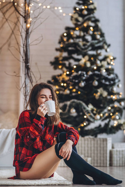 Pretty brunette girl sitting on the floor in red shirt and warm socks with cup of coffee and christmas tree behind - Foto, Imagem