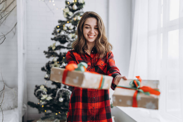 Christmas, New Year. Pretty woman in shirt and socks walking at home with christmas gift box, new year tree behin - 写真・画像