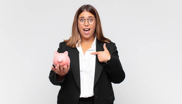 young woman with a piggy bank feeling happy, surprised and proud, pointing to self with an excited, amazed look - Foto, Bild