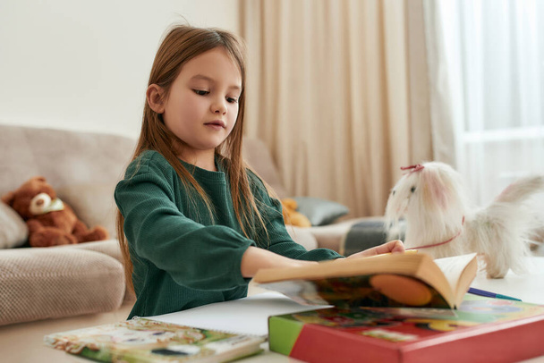Mała słodka dziewczyna siedzi przy stole w domu i czyta książkę w dużym pokoju - Zdjęcie, obraz