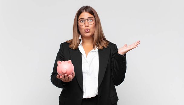 young woman with a piggy bank looking surprised and shocked, with jaw dropped holding an object with an open hand on the side - Photo, image