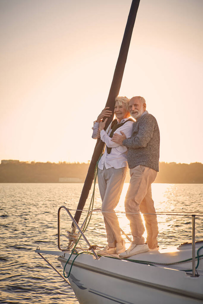 Full leghth of beautiful happy senior couple in love standing on the side of sail boat floating in sea at sunset, they are hugging and enjoying amazing evening view - Valokuva, kuva