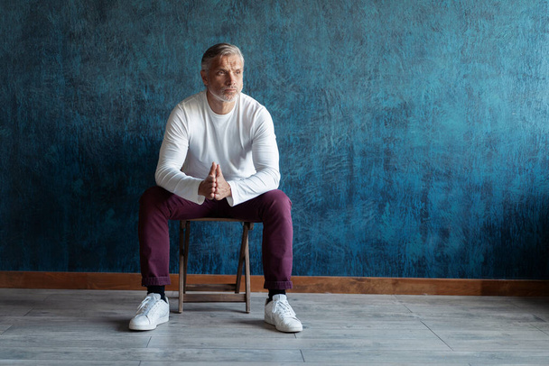 Portrait of a casual Grey-haired Mature handsome man sitting on a chair on dark blue background - Foto, afbeelding