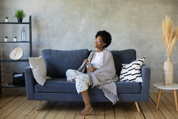 Feliz afroamericana joven mujer sentarse relajarse en acogedor sofá feliz de mudarse a un nuevo apartamento. Sonriente negro chica resto en cómodo sofá en sala de estar soñando - Foto, Imagen