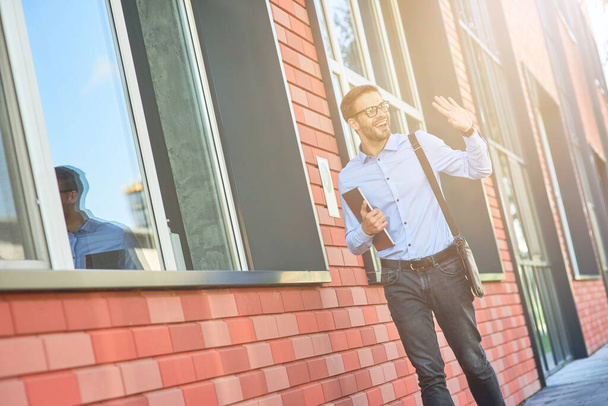 Een jonge blanke zakenman met een blauw hemd en een bril met z 'n laptop in z' n hand en zwaaiend, die hallo zegt terwijl hij buiten tegen kantoorgebouw staat. - Foto, afbeelding