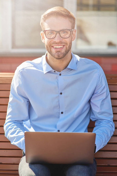 Pystysuora kuva nuoresta onnellisesta liikemiehestä, joka käyttää kannettavaa tietokonetta, katselee kameraa ja hymyilee istuessaan penkillä ulkona ja työskennellessään verkossa - Valokuva, kuva
