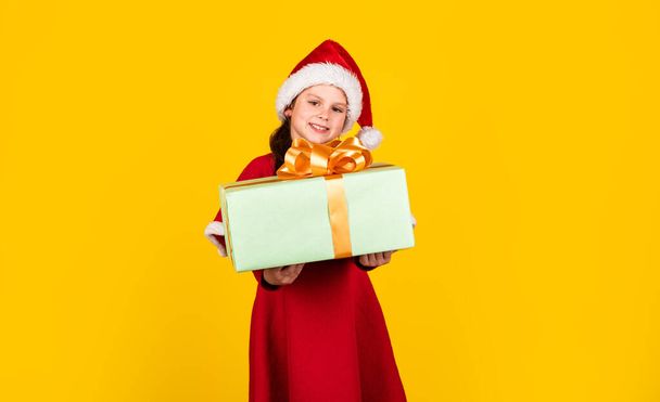 Pequeña niña Santa sombrero celebrar caja de regalo. Niño mantenga presente caja de fondo amarillo. Feliz Navidad y felices fiestas. Los sueños se hacen realidad. Vacaciones de invierno. Mi feliz día. Compras de regalos de Navidad. Paquete regalo - Foto, imagen