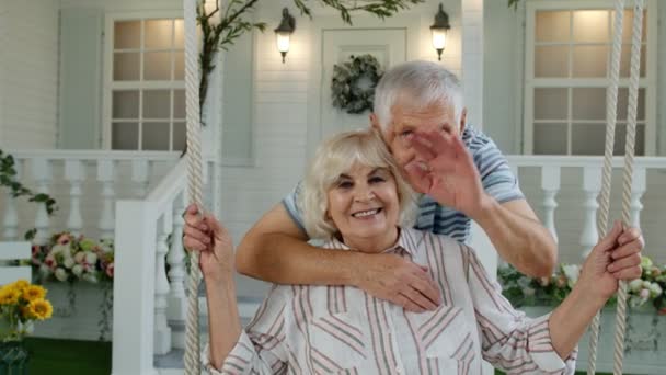 Ηλικιωμένο ζευγάρι στην μπροστινή αυλή στο σπίτι. Άντρας αγκαλιάζει γυναίκα. Ευτυχισμένη ώριμη οικογένεια χαιρετάει τα χέρια - Πλάνα, βίντεο