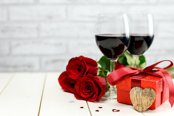 Η ιδέα του Αγίου Βαλεντίνου. Δύο ποτήρια κρασί, κόκκινα τριαντάφυλλα, κουτί δώρου, ξύλινη καρδιά και αναμμένα κεριά σε ένα λευκό ξύλινο τραπέζι. Επιλεκτική εστίαση. - Φωτογραφία, εικόνα