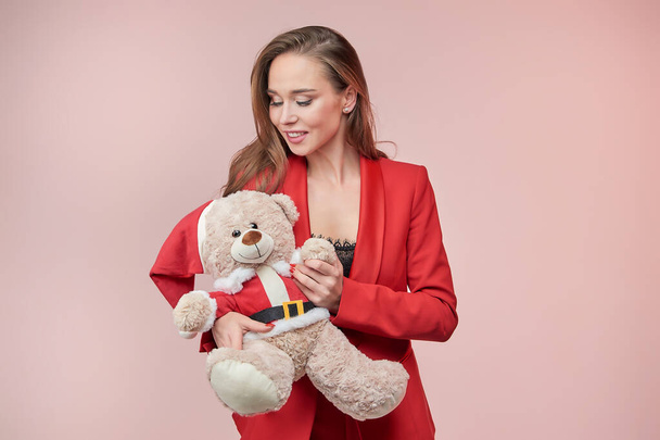 Smiling beautyful woman in a red jacket lovingly looks at a toy bear in the image of Santa Claus. New Year concept. - Photo, image