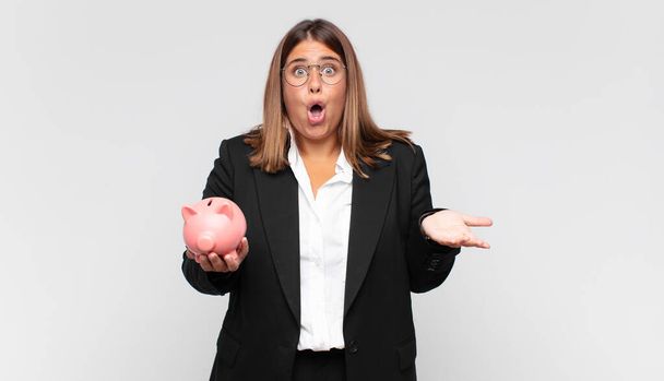 young woman with a piggy bank feeling extremely shocked and surprised, anxious and panicking, with a stressed and horrified look - Фото, изображение