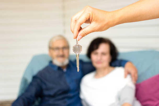 Realtor hand holding key to new house on blurred background of senior aged couple, sitting on sofa at meeting with real estate agent. Purchase or rent of apartment for retirement concept - Photo, Image