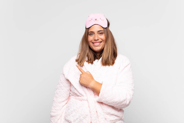 young woman wearing pajamas, smiling cheerfully, feeling happy and pointing to the side and upwards, showing object in copy space - Фото, зображення