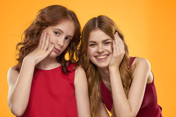 Egy vörös hajú lány és egy gyönyörű nő a sárga háttérben piros ruhában gesztikulálnak a kezükkel és a grimaszukkal. - Fotó, kép