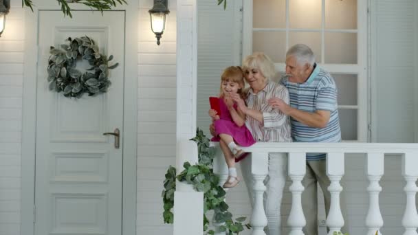 Starší pár sedí s vnučkou doma na verandě. Použití mobilního telefonu pro online videohovor - Záběry, video