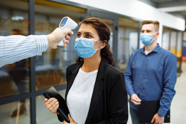 Büroangestellte werden während einer Coronavirus-Pandemie mit einem entfernten berührungslosen Thermometer überprüft. Junge Geschäftsfrau passiert Temperaturkontrolle auf dem Büroflur, um die Ausbreitung von Covid-19 zu verhindern - Foto, Bild