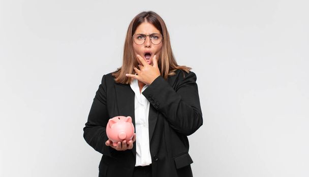 young woman with a piggy bank with mouth and eyes wide open and hand on chin, feeling unpleasantly shocked, saying what or wow - Foto, Imagem