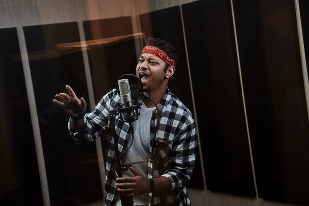 Retrato de un joven artista de hip hop enfocado, cantando en un micrófono de condensador mientras graba una canción en un estudio profesional - Foto, Imagen
