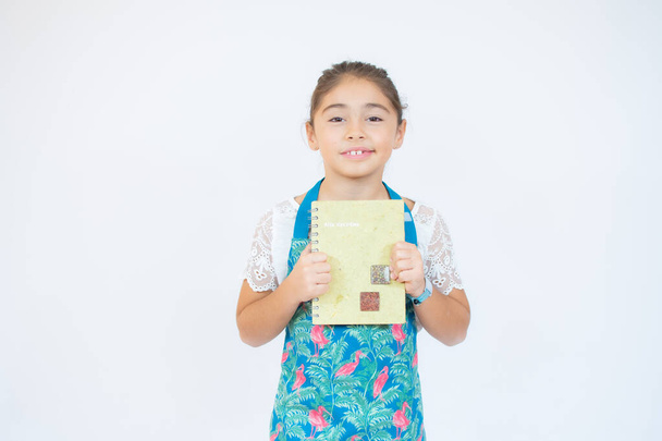 Portrait d'une petite fille dans la cuisine habillée en cuisinière souriante et tenant un livre de cuisine à la main. Concept de : nutrition, école de cuisine, éducation et famille. - Photo, image