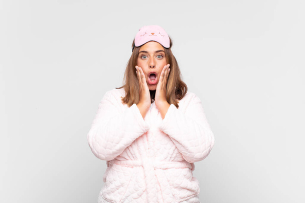 young woman wearing pajamas, feeling shocked and scared, looking terrified with open mouth and hands on cheeks - Foto, Imagem