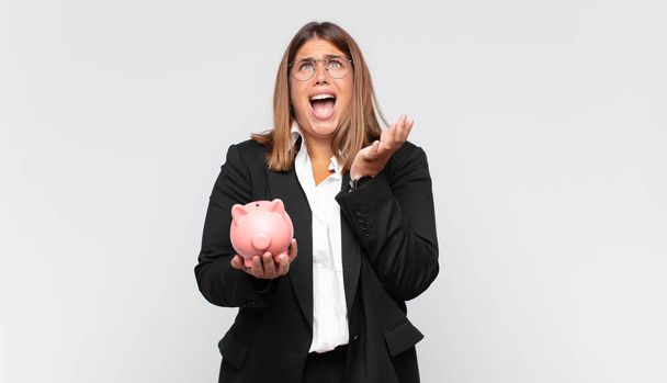 young woman with a piggy bank looking desperate and frustrated, stressed, unhappy and annoyed, shouting and screaming - 写真・画像
