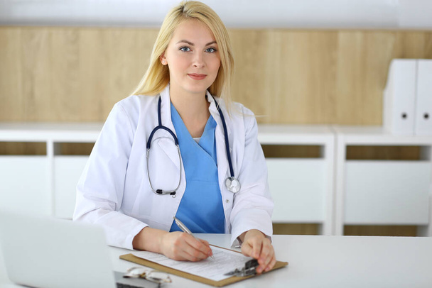 Доктор жінка на роботі, сидячи за столом у лікарні або клініці. Блондинка веселий лікар заповнює форму медичних записів. Дані та найкращий сервіс у медицині та охороні здоров'я
 - Фото, зображення