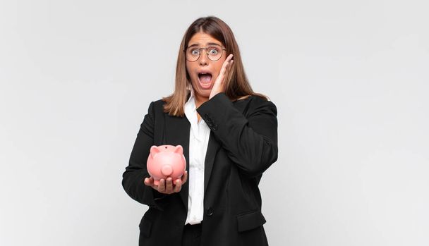 young woman with a piggy bank feeling happy, excited and surprised, looking to the side with both hands on face - Valokuva, kuva