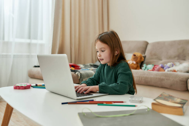 A small girl spending time alone in front of a laptop with her fluffy toys on a sofa during distance education - Foto, afbeelding