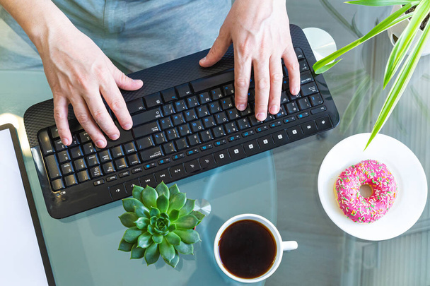 Male hands are typing on a wireless keyboard, succulent plant, a donut on a plate and a cup of coffee on a glass table. Home office, distance learning, remote work. Breakfast at workplace. Top view - Foto, afbeelding