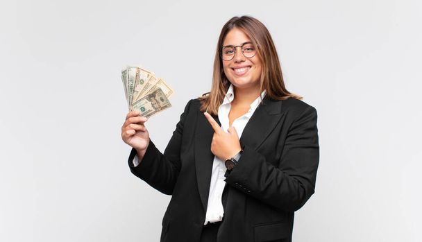 young woman with banknotes smiling cheerfully, feeling happy and pointing to the side and upwards, showing object in copy space - Fotografie, Obrázek