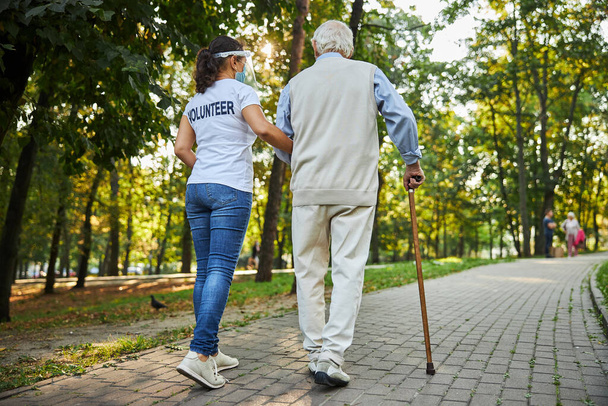 Alegre trabajadora social en camisa blanca caminando con anciano en la calle - Foto, imagen
