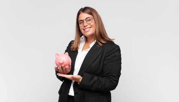 young woman with a piggy bank smiling cheerfully, feeling happy and showing a concept in copy space with palm of hand - Foto, imagen