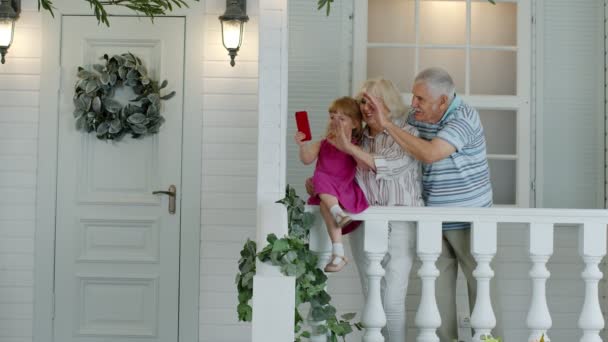 Az idősebbik otthon ül az unokájával a verandán. Mobiltelefon használata online videohíváshoz - Felvétel, videó