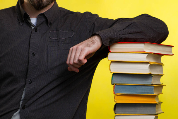 Έννοια της εκπαίδευσης. Ένας άντρας με μαύρο πουκάμισο ακουμπά σε μια στοίβα βιβλία. Κίτρινο φόντο. Κλείσε.. - Φωτογραφία, εικόνα