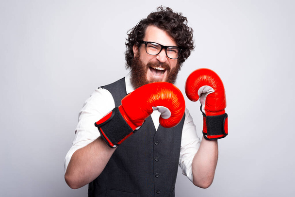 Γενειοφόρος νέος και χαρούμενος άντρας με γυαλιά και γάντια πυγμαχίας ουρλιάζει και κοιτάζει την κάμερα. - Φωτογραφία, εικόνα