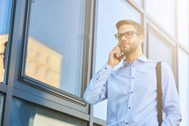 Joven guapo hombre de negocios caucásico en camisa azul hablando por teléfono mientras está de pie al aire libre - Foto, imagen