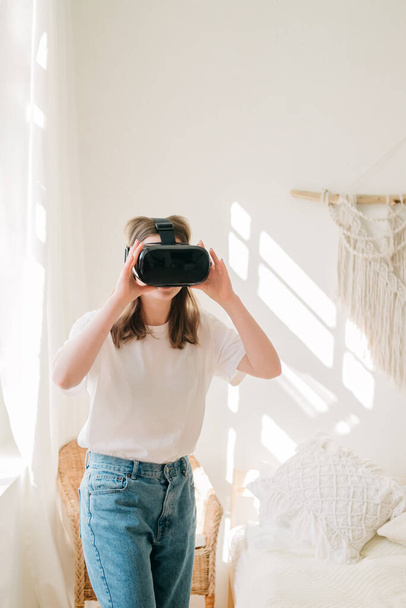 Young beautiful woman in white t-shirt and blue jeans plays game, dances with virtual reality headset helmet by the window in her room. Concept of entertainment, learning at home during quarantine. - Fotografie, Obrázek